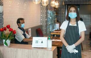 asiático camarera mujer vistiendo cara mascaras y participación un infrarrojo frente termómetro a cheque cuerpo temperatura para virus síntomas de clientes antes de entrando el restaurante café tienda. foto