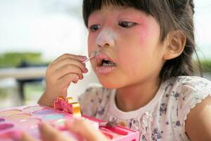 adorable pequeño niño asiático niña pinturas su boca con rosado niños cabezas y mira en el espejo. un niño obras de teatro a hogar en un juguete belleza salón. incrementar aprendizaje desarrollo para preescolares foto