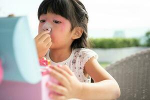 Adorable little child Asian girl paints her mouth with pink children heads and looks in the mirror. A child plays at home in a toy beauty salon. Increase learning development for preschoolers. photo