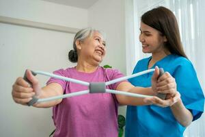 Happy elderly female exercising with rubber expander with Young physiotherapist for recovery arm muscles in a physiotherapy center. Concept of happy retirement with care from a caregiver and health. photo