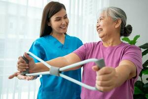 Happy elderly female exercising with rubber expander with Young physiotherapist for recovery arm muscles in a physiotherapy center. Concept of happy retirement with care from a caregiver and health. photo