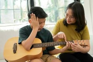 Asian mother embraces son, Asian boy playing guitar and mother embrace on the sofa and feel appreciated and encouraged. Concept of a happy family, learning and fun lifestyle, love family ties photo