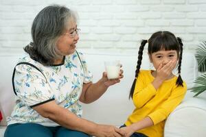 Happy elderly Asian Grandma sits beside her granddaughter and feeds fresh milk from glass for breakfast at home. Concept of a happy family and takes care together, preschool health care photo