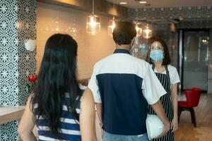 asiático camarera mujer vistiendo cara mascaras y participación un infrarrojo frente termómetro a cheque cuerpo temperatura para virus síntomas de clientes antes de entrando el restaurante café tienda. foto