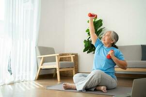 anciana asiática levantando pesas para hacer ejercicio y hacer ejercicio en casa. mujer madura activa haciendo ejercicio de estiramiento en la sala de estar. ejercicio activo y saludable para personas mayores, mayores y mayores. foto