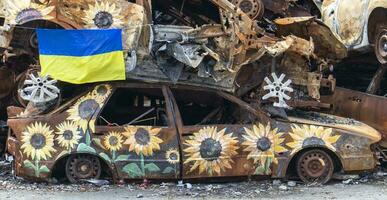 The national flag of Ukraine against the background of ruins and rusty metal of destroyed cars. A blue-yellow flag waving in the wind under the sun in a ruined city. Ukraine, Kyiv - April 29, 2023. photo