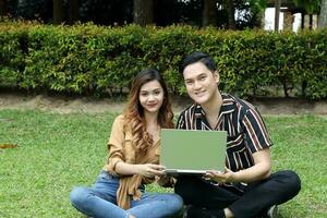 Young Asian Malay man woman outdoor green park sit on grass use study discuss laptop look at camera photo