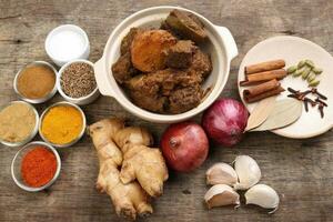 Beef bhuna dry curry with potato in ceramic bowl ingredients chilli cumin coriander turmeric powered salt onion garlic ginger bay leaf cinnamon cardamom clove over rustic wood background photo