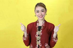 Asian woman traditional red maroon kebaya sharong on yellow paper background posing thumbs up photo