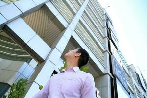 joven asiático medio oriental hombre en frente de un alto edificio Mira arriba pensar sueño foto