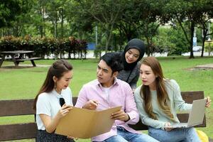 Young asian malay chinese man woman outdoor on park bench study talk discuss point laptop file book photo