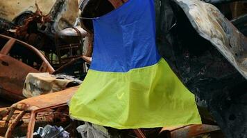 das National Flagge von Ukraine gegen das Hintergrund von Ruinen und rostig Metall von zerstört Autos. Krieg im Ukraine. ein Blau Gelb Flagge winken im das Wind unter das Sonne im ein bombardiert Stadt. video