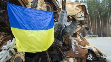 The national flag of Ukraine against the background of ruins and rusty metal of destroyed cars. War in Ukraine. A blue-yellow flag waving in the wind under the sun in a bomb-ravaged city. photo