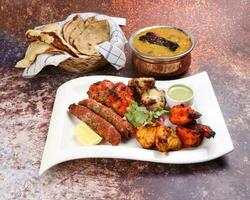 Mixed grill kebab chicken mutton beef prawn fish mint sauce lemon pickled onion bread basket chapati naan tandoori paratha dal lentil fry tadka copper pot on white dish over rustic background photo