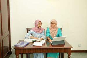 Two young Asian Malay Muslim woman wearing headscarf at home office student sitting at table talk mingle look at computer book document study discuss smile happy photo