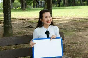 Young asian chinese woman outdoor on park bench hold whiteboard copy text space smile look point photo