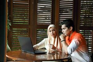 Young asian malay man woman with headscarf sit at rustic cafe table talk discuss business study read laptop computer photo