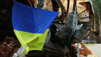 de nationaal vlag van Oekraïne tegen de achtergrond van ruïnes en roestig metaal van vernietigd auto's. oorlog in Oekraïne. een blauw Geel vlag golvend in de wind onder de zon in een bom verwoest stad. video