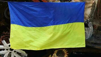 The national flag of Ukraine against the background of ruins and rusty metal of destroyed cars. War in Ukraine. A blue-yellow flag waving in the wind under the sun in a bomb-ravaged city. video