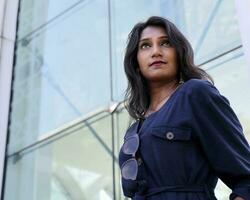 Young attractive beautiful Indian woman in dark blue formal jumpsuit look forward confidently hands in pocket front of a tall glass urban building photo