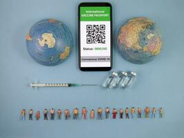 Miniature human figure figurine male female doll row vaccine passport digital paper book bottle medical injection syringe needle world map globe border copy text sign space on blue paper background photo