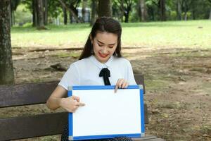 joven asiático chino mujer al aire libre en parque banco sostener pizarron Copiar texto espacio sonrisa Mira punto foto