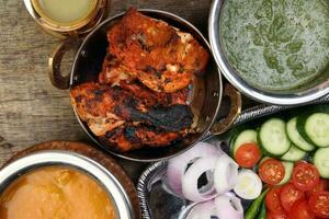 Spicy red Chicken grilled tikka tandoori nan bread green spinach curry yogurt sauce dal tomato cucumber onion salad set in metal stainless steel brass copper pot on rustic vintage wood background photo