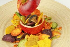 Healthy vegetable chips yellow sweet potato purple carrot green radish green beans and shiitake mushrooms in hollow red apple white background wooden plate photo