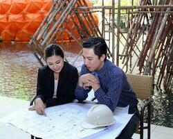 South East Asian young Malay Chinese man woman formalwear sit at table construction site study look plan blueprint photo