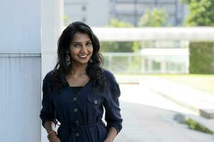 Young attractive beautiful Indian woman in dark blue formal jumpsuit look forward confidently leaning on urban building wall look at camera smiling photo