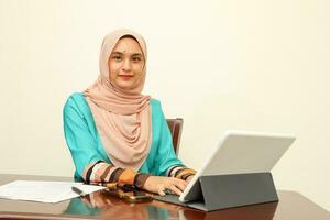 joven asiático malayo musulmán mujer vistiendo Pañuelo a hogar oficina estudiante sentado a mesa hablar mezclarse Mira a computadora pan de maíz libro documento estudiar discutir hablar sonrisa contento Mira a cámara tipo foto