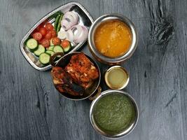 Spicy red Chicken grilled tikka tandoori nan bread green spinach curry yogurt sauce dal tomato cucumber onion salad set in metal stainless steel brass copper pot on black char wood background photo