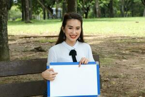 Young asian chinese woman outdoor on park bench hold whiteboard copy text space smile look point photo