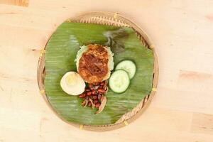 Nasi Lemak fragrant rice cooked in coconut milk served with sambal friend peanut anchovy boiled egg packed in banana leaf on round bamboo plate on wooden background photo