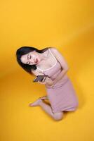 Beautiful young south east Asian woman sit down on floor yellow orange color background hold use text smart phone pose fashion style elegant beauty mood photo