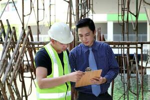 sur este asiático joven malayo chino hombre vistiendo blanco amarillo la seguridad casco chaleco Corbata estudiar ver hablar portapapeles al aire libre construcción sitio foto