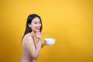 Beautiful young south east Asian woman holding chines empty chopstick Chinese soup spoon bowl utensil pretend acting posing see eat taste smell feed offer satisfaction yummy yellow orange background photo