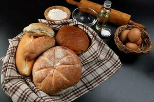 Freshly baked bread loaf bun roll round long mix verity wrapped in checkered kitchen fabric napkin towel wheat flower oil water salt eggs rolling pin slate stone over black background photo