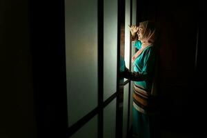 young Asian Malay muslim woman wearing headscarf baju kurung dress at home in front window mood light on face dark background peaceful content calm tranquil break trough wonder photo