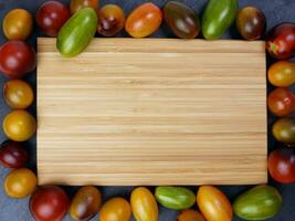 Red orange yellow green tomato mix variety on around wooden chopping board frame copy text space over black slate stone background photo