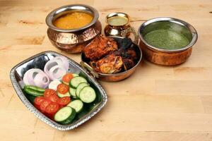 Spicy red Chicken grilled tikka tandoori nan bread green spinach curry yogurt sauce dal tomato cucumber onion salad set in metal stainless steel brass copper pot on light wood background photo