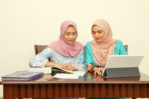 Two young Asian Malay Muslim woman wearing headscarf at home office student sitting at table talk mingle look at computer book document study discuss smile happy photo