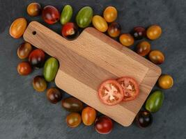 Red orange yellow green tomato mix variety on around wooden chopping board frame copy text space over black slate stone background photo