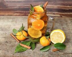 Liquid ice lemon orange tea with slice green leaf cinnamon stick in transparent glass jar mug on rustic wood background whole fruit photo