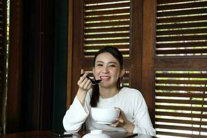 Young asian woman sit at rustic cafe eat from chinese spoon from soup noodle from bowl look front at camera photo