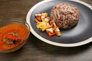 Nasi dagang special brown white rice cooked with coconut milk pickled fruit vegetable pineapple carrot on black off white rim plate fish curry in bowl over rustic dark wooden background photo