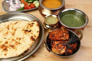 Spicy red Chicken grilled tikka tandoori nan bread green spinach curry yogurt sauce dal tomato cucumber onion salad set in metal stainless steel brass copper pot on light wood background photo