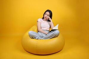 Beautiful young south east Asian woman sits on a yellow beanbag seat orange yellow color background pose fashion style elegant beauty mood expression rest relax read book think photo