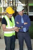 sur este asiático joven malayo chino hombre vistiendo blanco amarillo la seguridad casco chaleco Corbata estudiar ver hablar portapapeles al aire libre construcción sitio foto