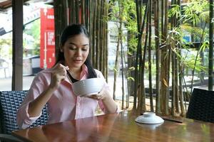 Young asian malay woman sit at rustic cafe table eat with chinese spoon from soup noodle bowl look down at food photo